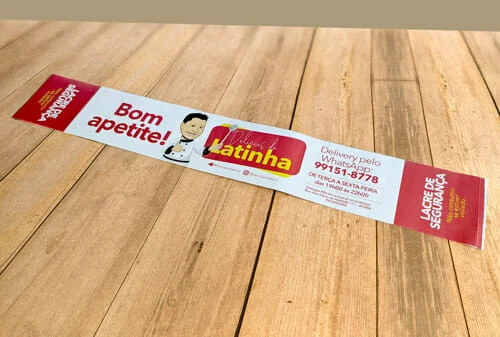 Rótulo Adesivo Personalizado da Marca "Delícias da Latinha" tem o desenho de um homenzinho de roupa de cozinheiro e uma colher na mão, também está escrito "bom apetite" e "lacre de segurança" no rótulo. está sobre uma mesa de madeira.