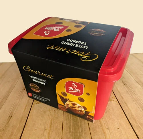 Envoltório de sorvete personalizado da Empresa Maipu, o envoltório é preto, escrito "Gourmet" em dourado, e abaixo do gourmet o sabor do sorvete que é "leite ninho trufado", tem um retângulo arrendondado no canto inferior direito, com o logotipo da Maipu, e abaixo uma bola de sorvete amarela com gotas de chocolate, a bola está caindo dentro de uma calda de chocolate, que está esparramando para cima, na parte superior também tem a mesma foto. A embalagem de sorvete rodeada com o envoltório está sobre uma mesa de madeira.