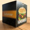 caixa Hamburguer Personalizada, a cor predominante é preta, com uma textura de hambúrgueres desenhados em amarelo. Na face a esquerda é possível ver uma tira em amarela com o escrito 