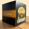 caixa Hamburguer Personalizada, a cor predominante é preta, com uma textura de hambúrgueres desenhados em amarelo. Na face a esquerda é possível ver uma tira em amarela com o escrito "saboroso" em branco e na face a direita uma foto de um lanche com a palavra "hambúrguer" abaixo. A caixa está sobre uma mesa de madeira.