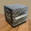 Caixa hambúrguer personalizada na cor preta, com uma textura de fundo branca com vários desenhos de sanduíches, cachorro-quentes, refrigerantes e batatas. É possível ver duas faces da embalagem, sendo a face esquerda com o desenho de um sanduíche grande, sobre fundo preto e na lateral direita, a palavra "hambúrguer" dentro da silhueta de um hambúrguer.