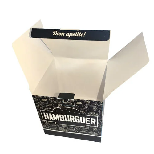 Caixa hambúrguer personalizada na cor preta, com uma textura de fundo branca com vários desenhos de sanduíches, cachorro-quentes, refrigerantes e batatas. Na parte da frente é possível ver palavra "hambúrguer" dentro da silhueta de um hambúrguer. A embalagem está aberta e na lingueta que fecha a caixa está escrito "Bom apetite".