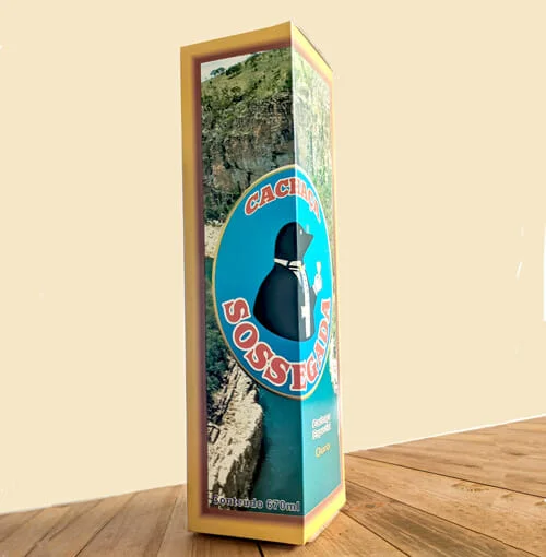 Caixa de bebida personalizada da "Cachaça Sossegada", atrás do logotipo tem uma foto de um cânion com água bem azul, ao redor da imagem tem uma borda marrom e amarela. A caixa está sobre uma mesa de madeira.
