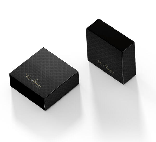 Duas caixas cinza chumbo com arabescos pretos, ela se assemelha a uma caixa de fósforos e tem um logotipo escrito a mão em dourado "Tati Negrão Semijoias". Uma está em na vertical e a outra na horizontal.