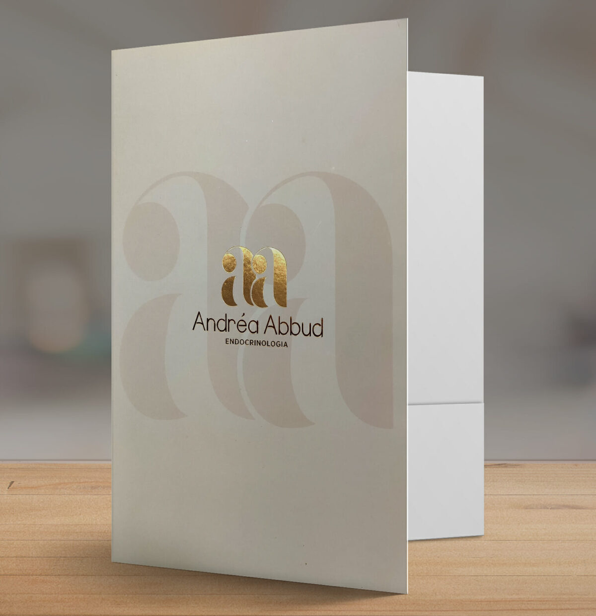 Pasta Ultra Luxo, com hot stamping, laminação fosca e alto relevo, com o logo AA Andrea Abud, na parte da frente. Ela está aberta sobre uma mesa de madeira.