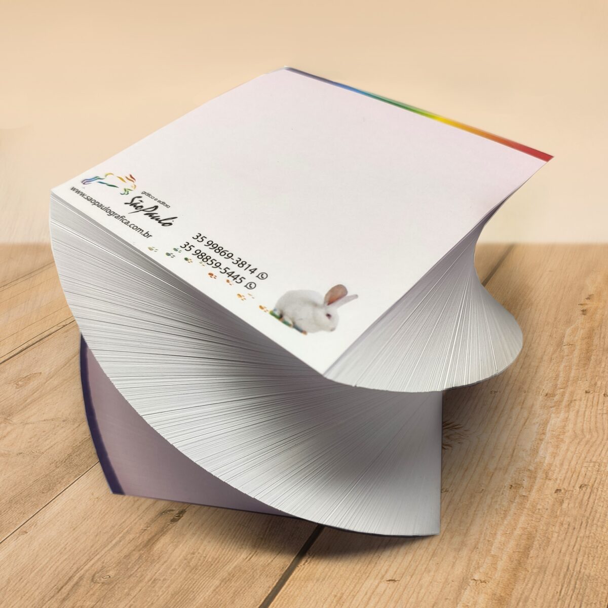 Bloco (500 folhas) personalizado com o seu logotipo, em papel sulfite 56g. É possível ver o bloquinho de forma torcida sobre uma mesa de madeira.
